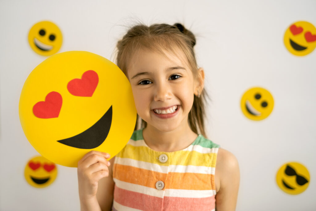A girl holding a paper emoji upon active listening at an advanced level. She chose a smiling emoji with hearts for eyes.